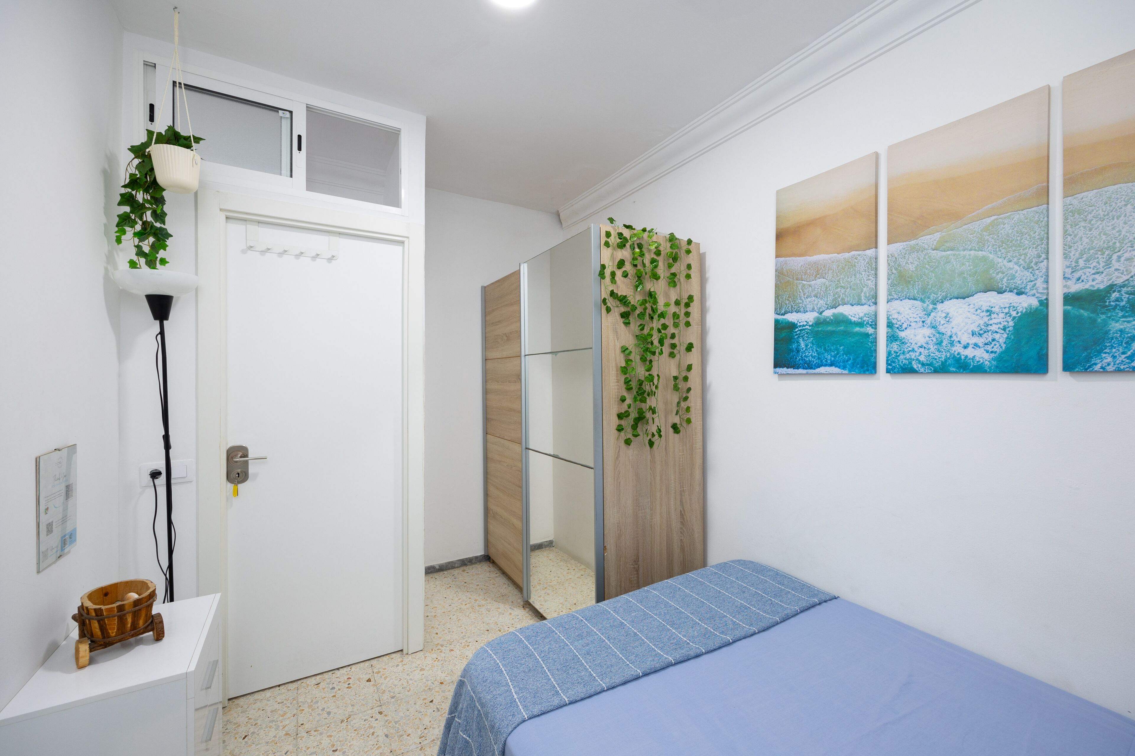 Un dormitorio con cama, armario blanco, mesita con cesta, planta en la pared y una pintura de olas.