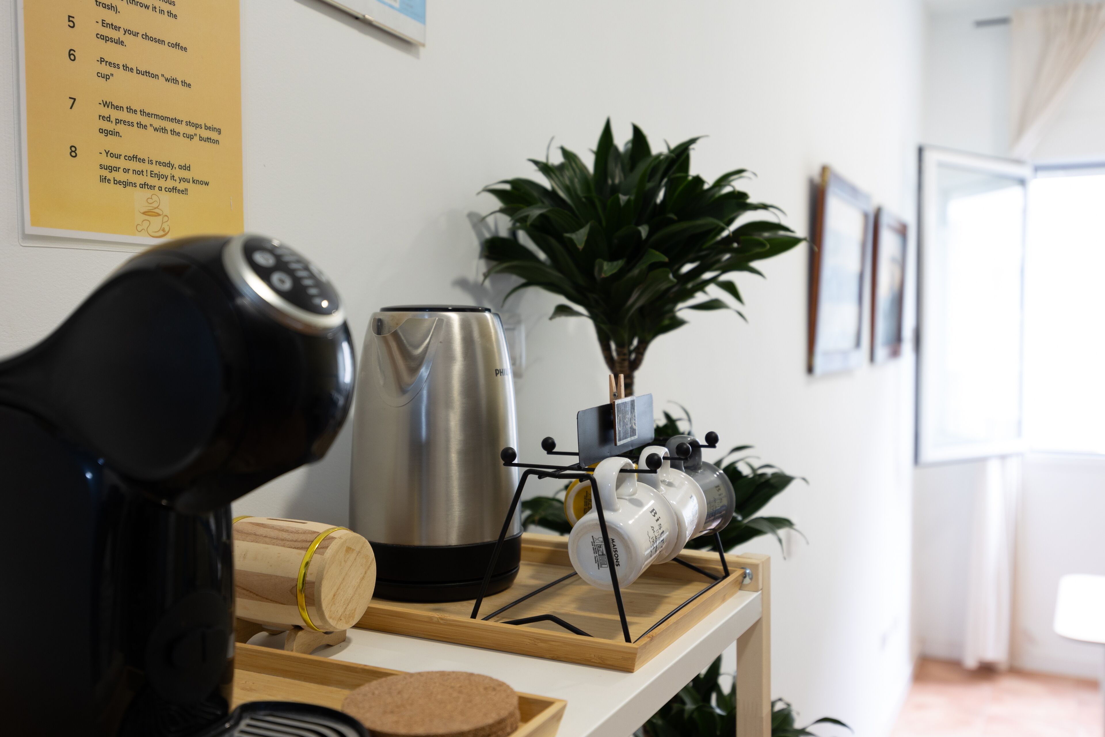 Una cafetera, un hervidor y una bandeja con tazas sobre una mesa de madera en una habitación con una planta y cuadros en la pared.