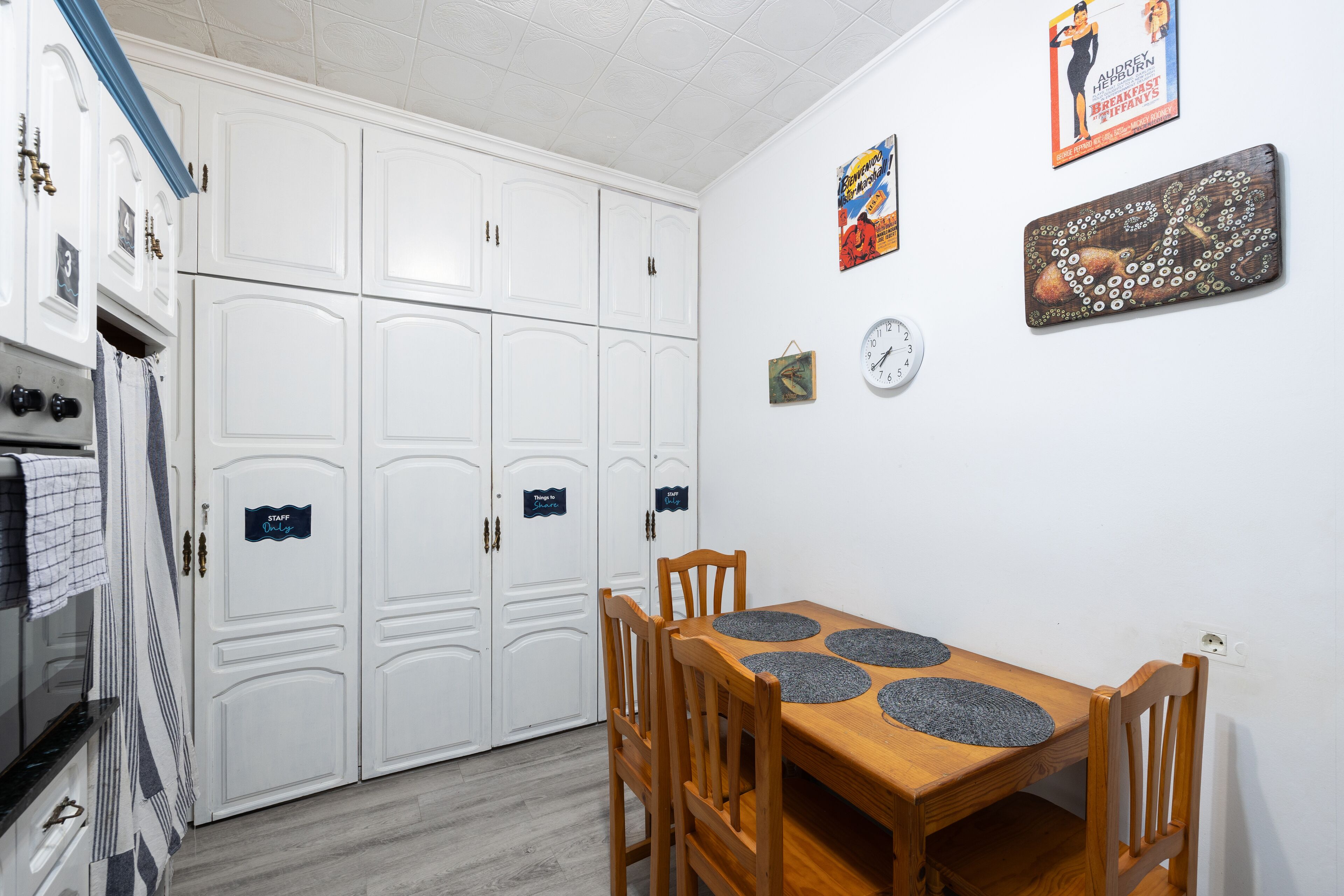 A dining area with a wooden table and chairs, a clock, and a wall with various items including a poster and a framed picture.
