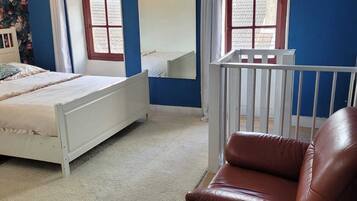 A bedroom with a bed, a white crib, a brown leather chair, and two windows with curtains.