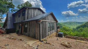 A rustic wooden cabin with a wooden deck, surrounded by a stone wall and gravel, overlooking a lush green valley. - Estill Oaks - Luxe Mountain View Retreat -Hot Tub - Secluded 80-Acre Escape for 16 Guests (Estill County)
