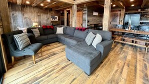 A rustic interior with a large L-shaped sectional sofa, wooden flooring, and exposed beams. - Estill Oaks - Luxe Mountain View Retreat -Hot Tub - Secluded 80-Acre Escape for 16 Guests (Estill County)