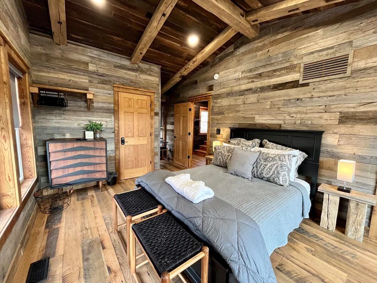 Una habitación rústica de madera con una cama grande, un banco, un aparador y una mesita de noche con una lámpara.