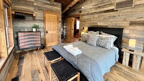 A rustic wooden room with a large bed, a bench, a dresser, and a small table with a lamp. - Estill Oaks - Luxe Mountain View Retreat -Hot Tub - Secluded 80-Acre Escape for 16 Guests (Estill County)
