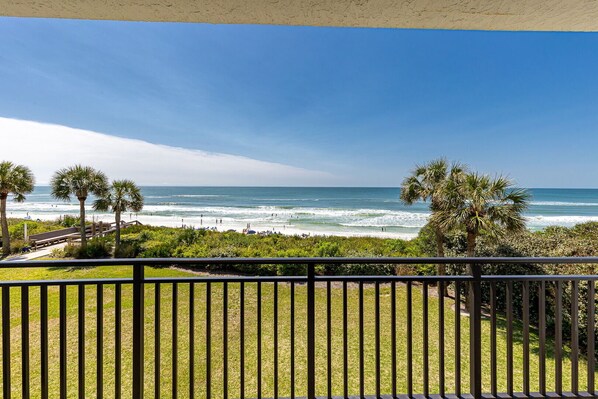 A balcony with a view of the ocean, palm trees, and a wooden walkway leading to the beach.