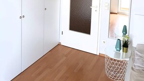 Ein Zimmer mit Schrank, einer Tür mit strukturierter Verkleidung, einem runden Tisch mit Vase und Pflanze sowie einem Ausblick durch ein Fenster.