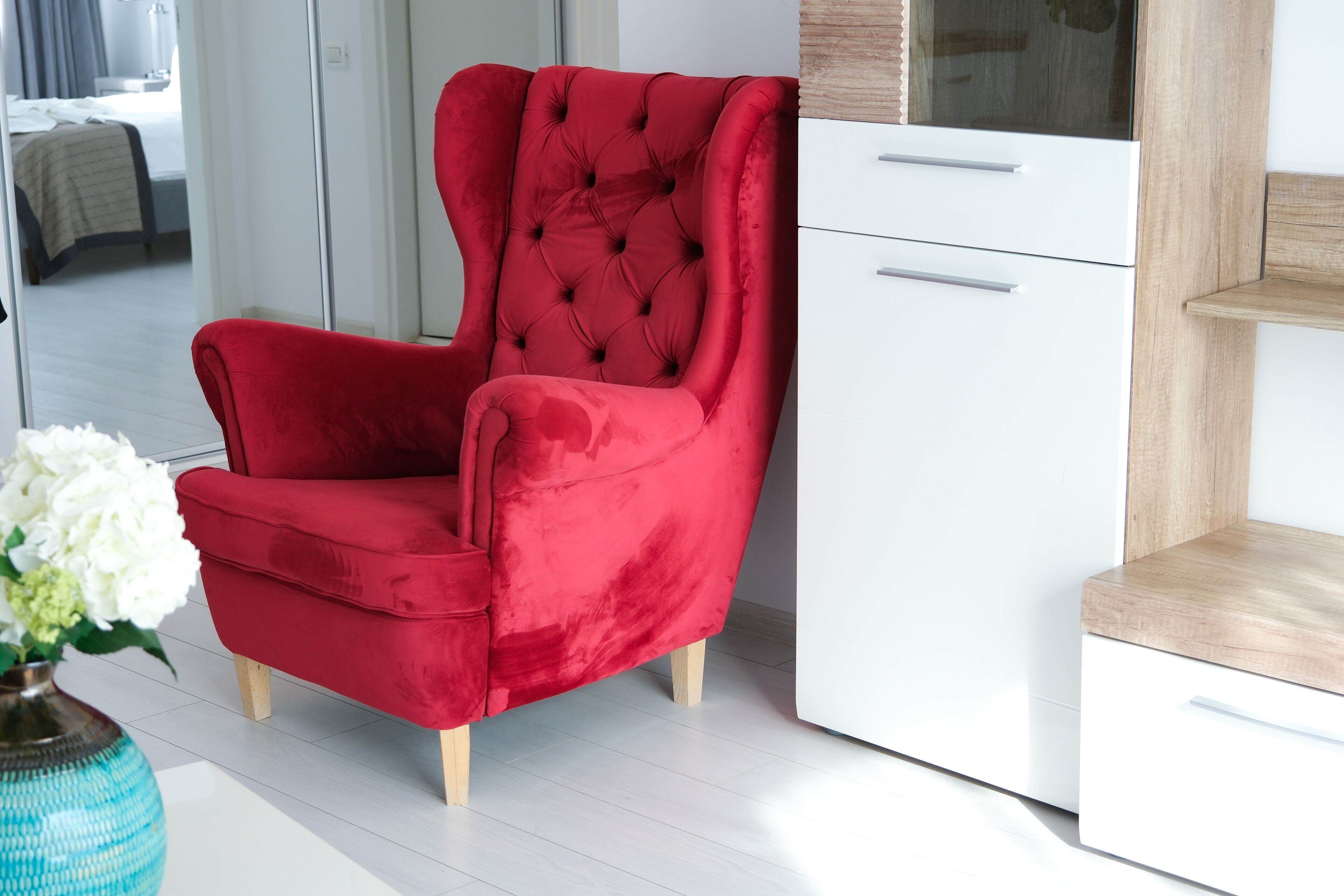 A red velvet armchair with wooden legs, a white dresser, and a wooden shelf unit in a modern room with white flooring.