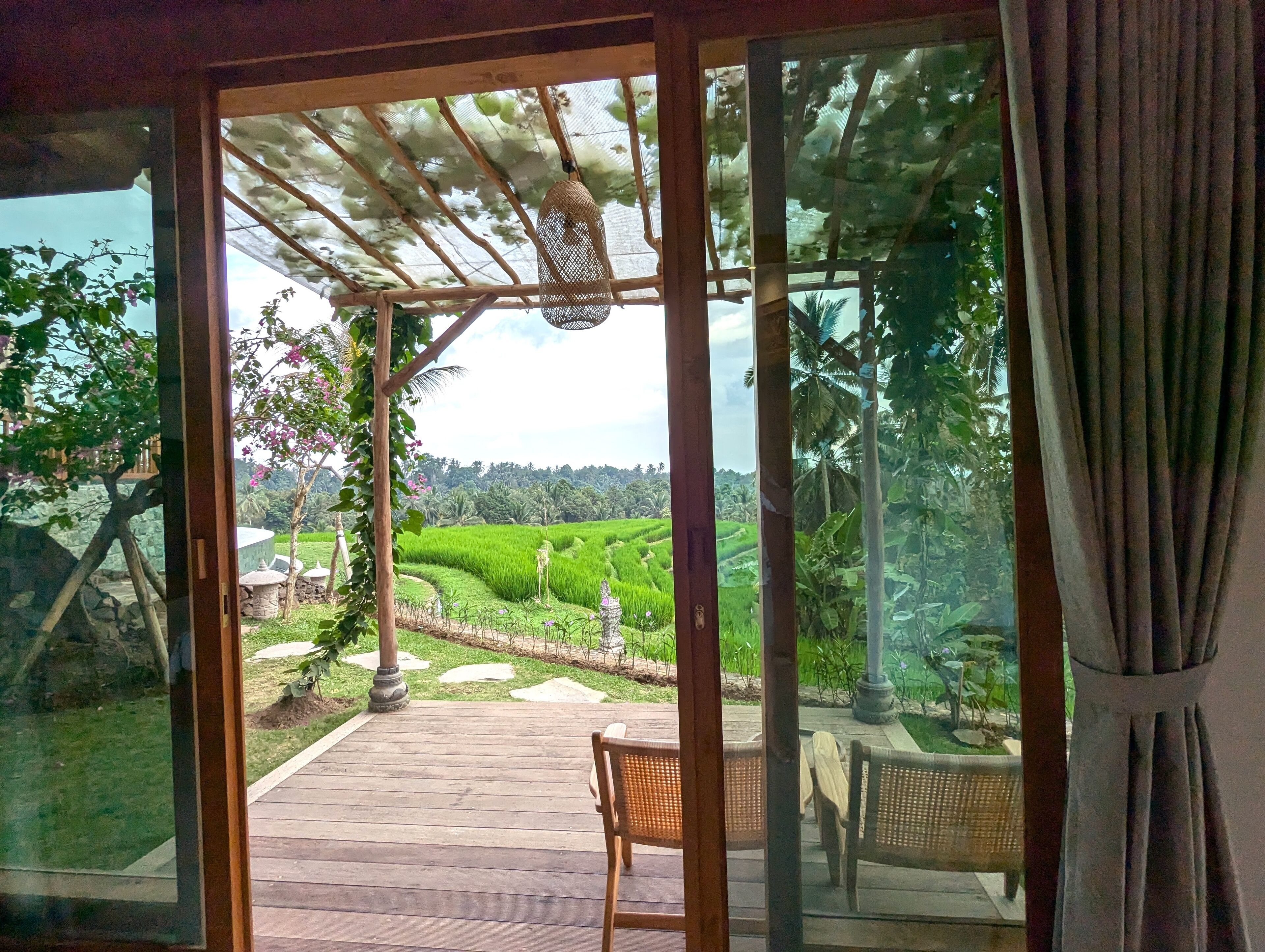 A wooden deck with a chair, a pergola with a hanging basket, and a view of a lush green landscape with terraced fields.