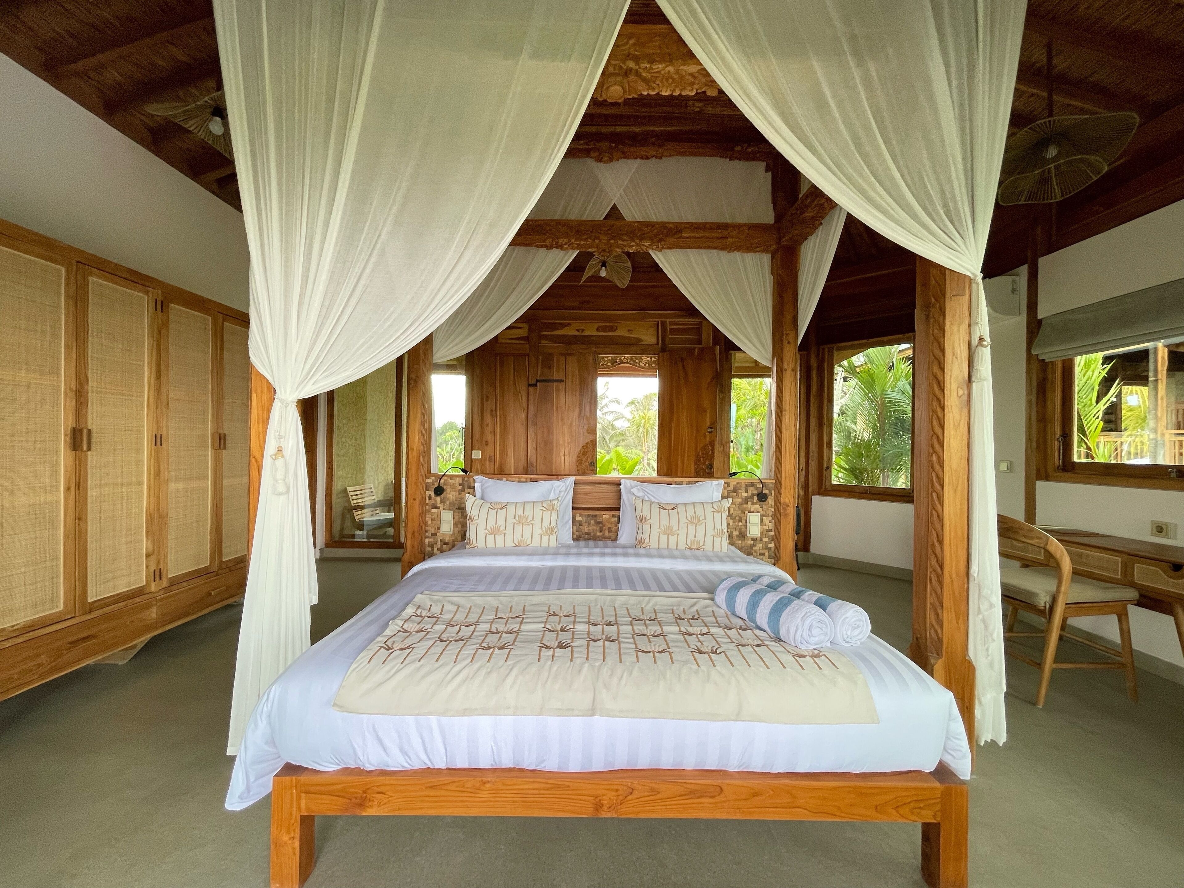 A wooden four-poster bed with white bedding and a canopy, a wooden desk, and a view of greenery outside.