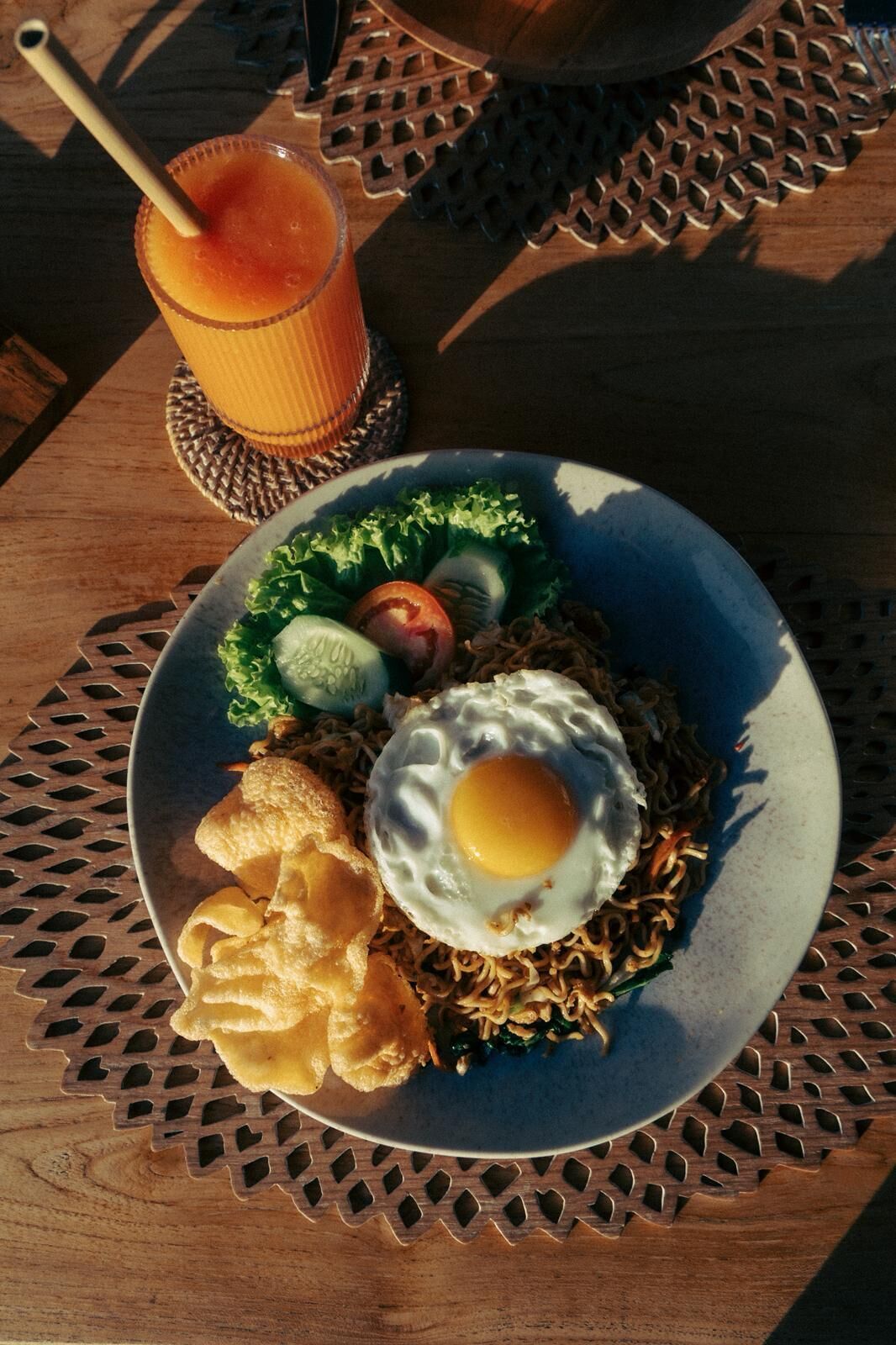 A plate of food with noodles, vegetables, and a fried egg, accompanied by a drink and chips on a wooden table.