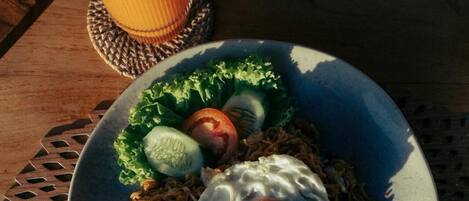 A plate of food with noodles, vegetables, and a fried egg, accompanied by a drink and chips on a wooden table.