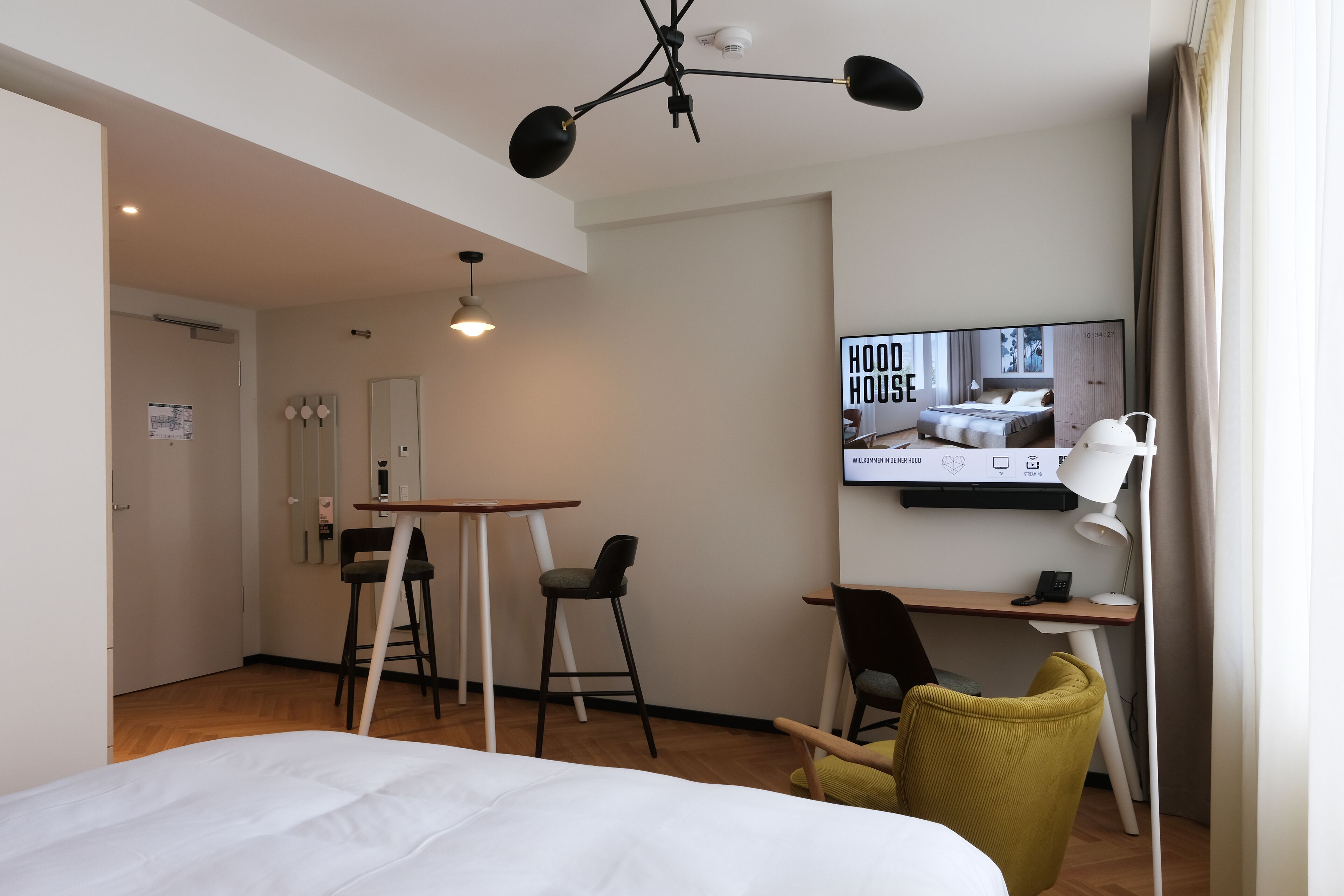A modern hotel room with a bed, a desk, a chair, and a television displaying a room.