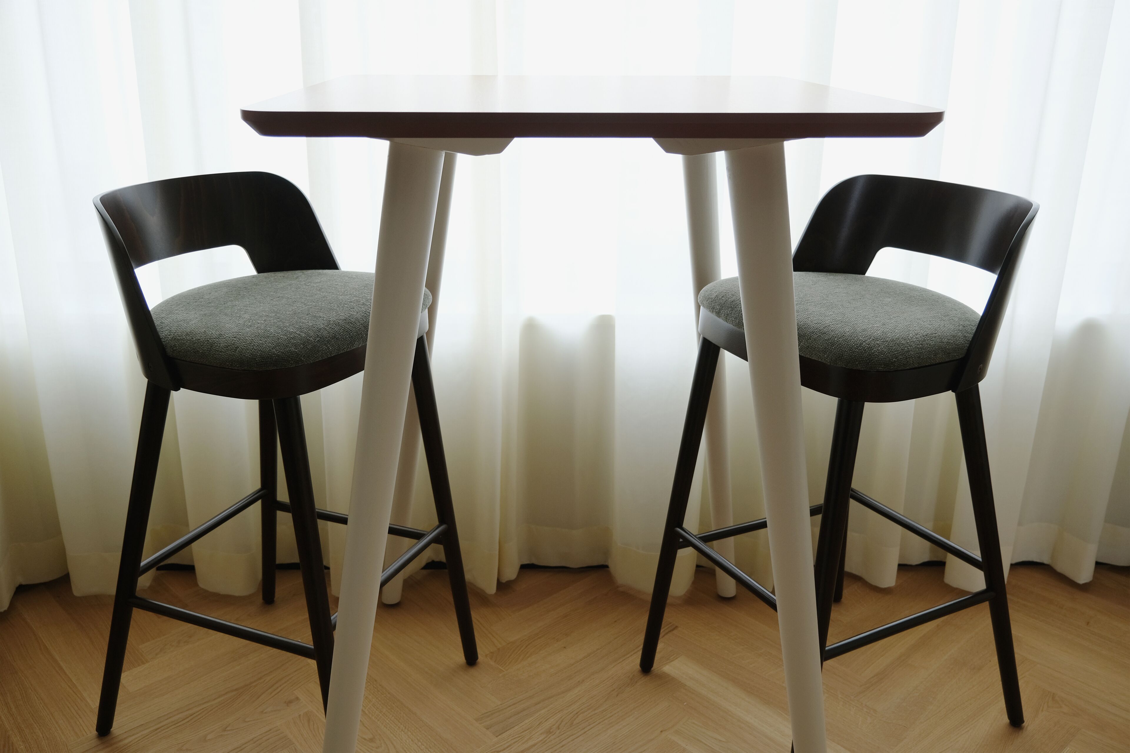 Deux chaises noires avec des coussins gris et une table blanche aux pieds noirs, dans une pièce dont le sol est recouvert de parquet et où sont accrochées des rideaux blancs.