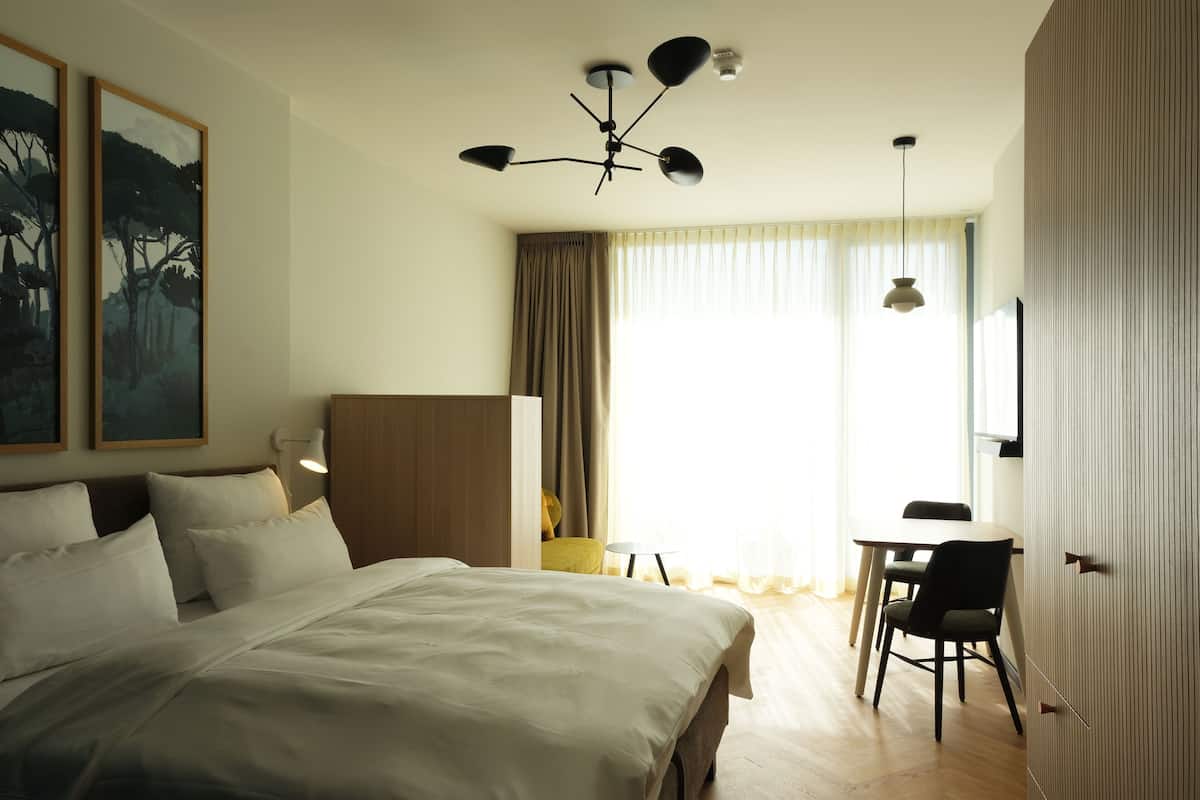 A hotel room with a large bed, a desk, and a chair. There is a ceiling fan, a window with curtains, and a painting on the wall.