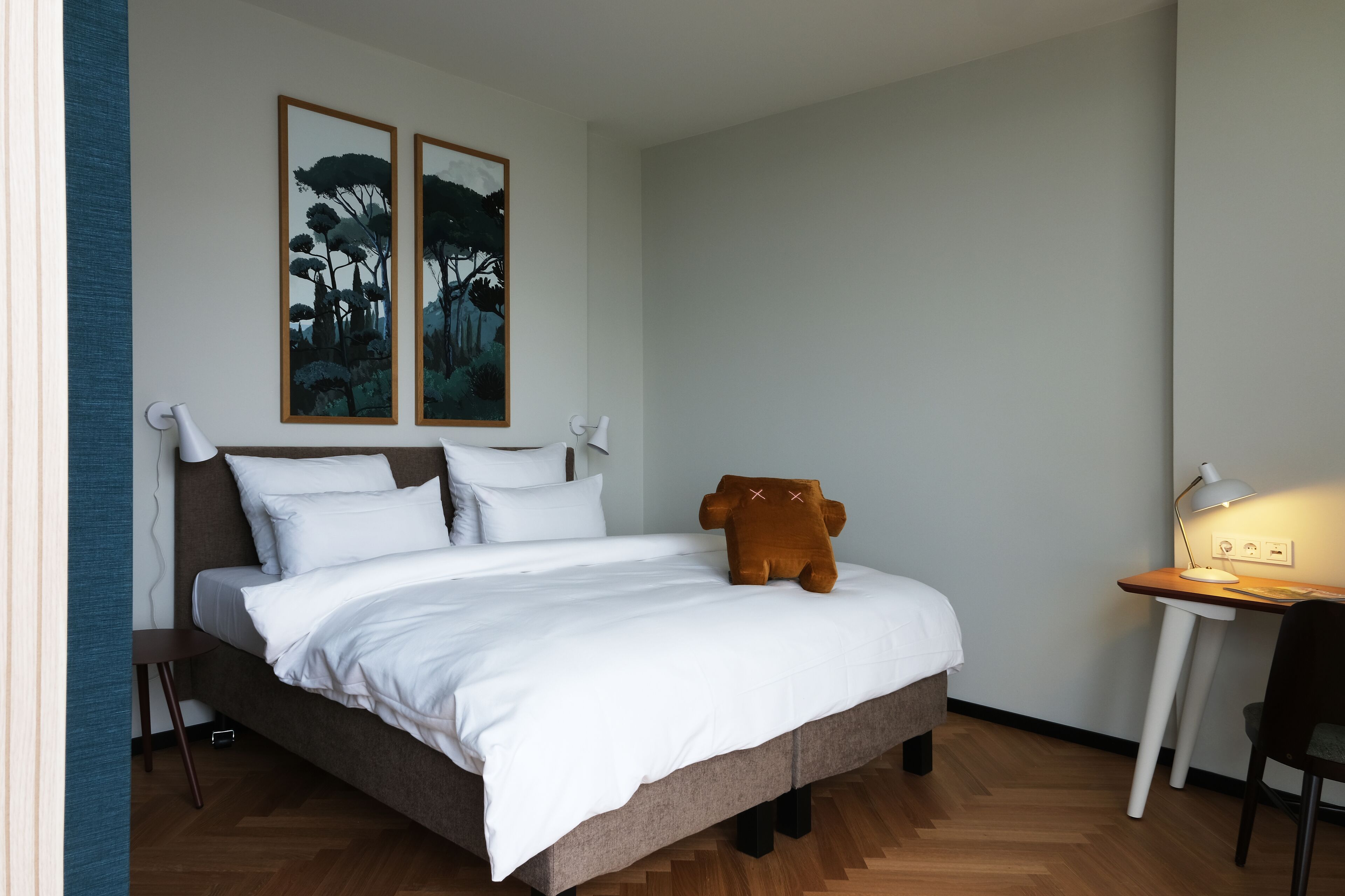 Ein ordentlich gemachtes Bett mit weißer Bettwäsche, einem kissenförmigen braunen Bären und zwei gerahmten Bildern an der Wand.