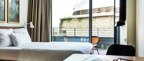 A hotel room with a bed, a table with glasses, and a view of a building through large windows.