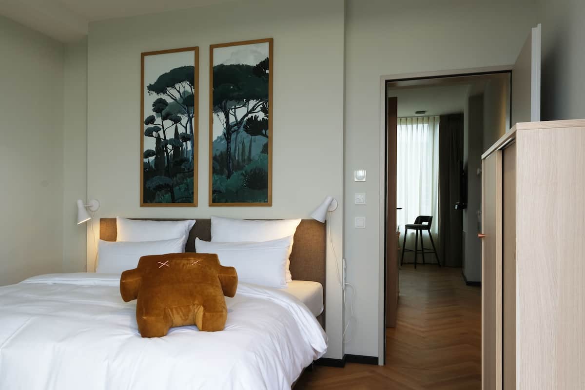 A hotel room with a bed, a brown teddy bear on the bed, two paintings on the wall, and a wooden wardrobe.