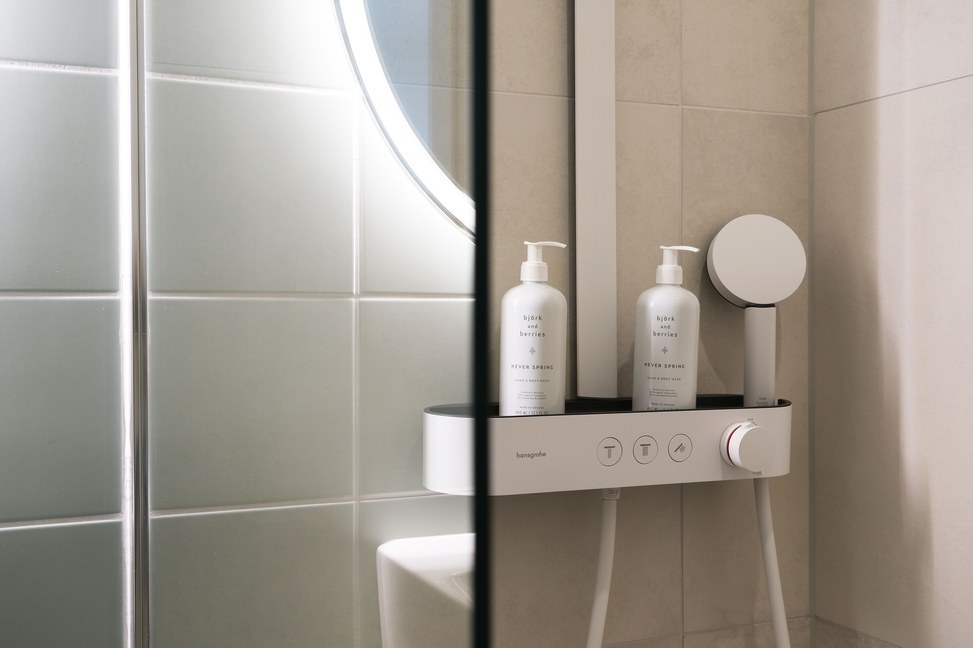 A modern bathroom with a glass shower enclosure, a white sink, and two bottles of 'bjark and berries' shampoo and conditioner.