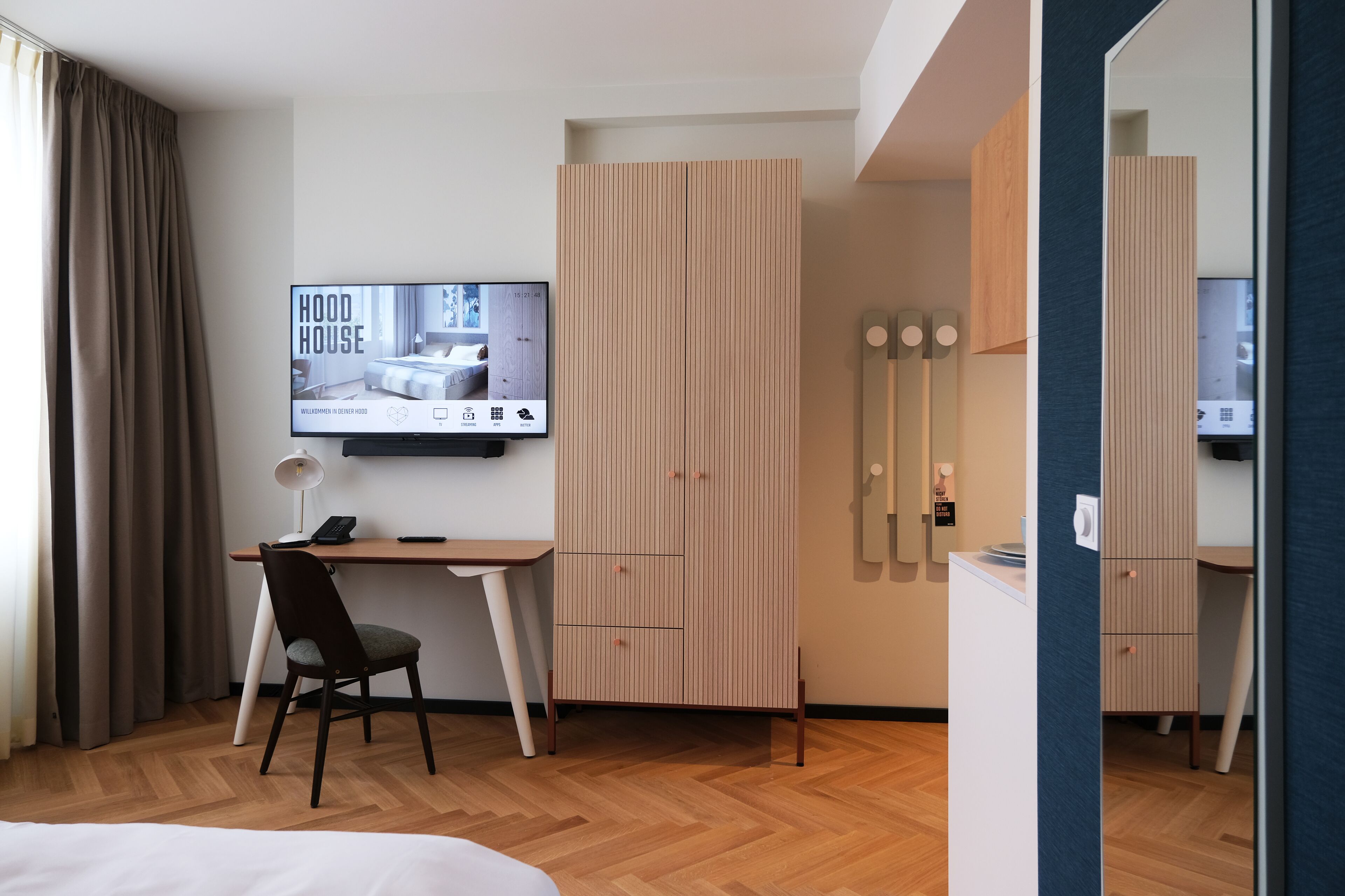 A modern hotel room with a wooden desk, a chair, a television mounted on the wall, and a wardrobe with vertical slats.