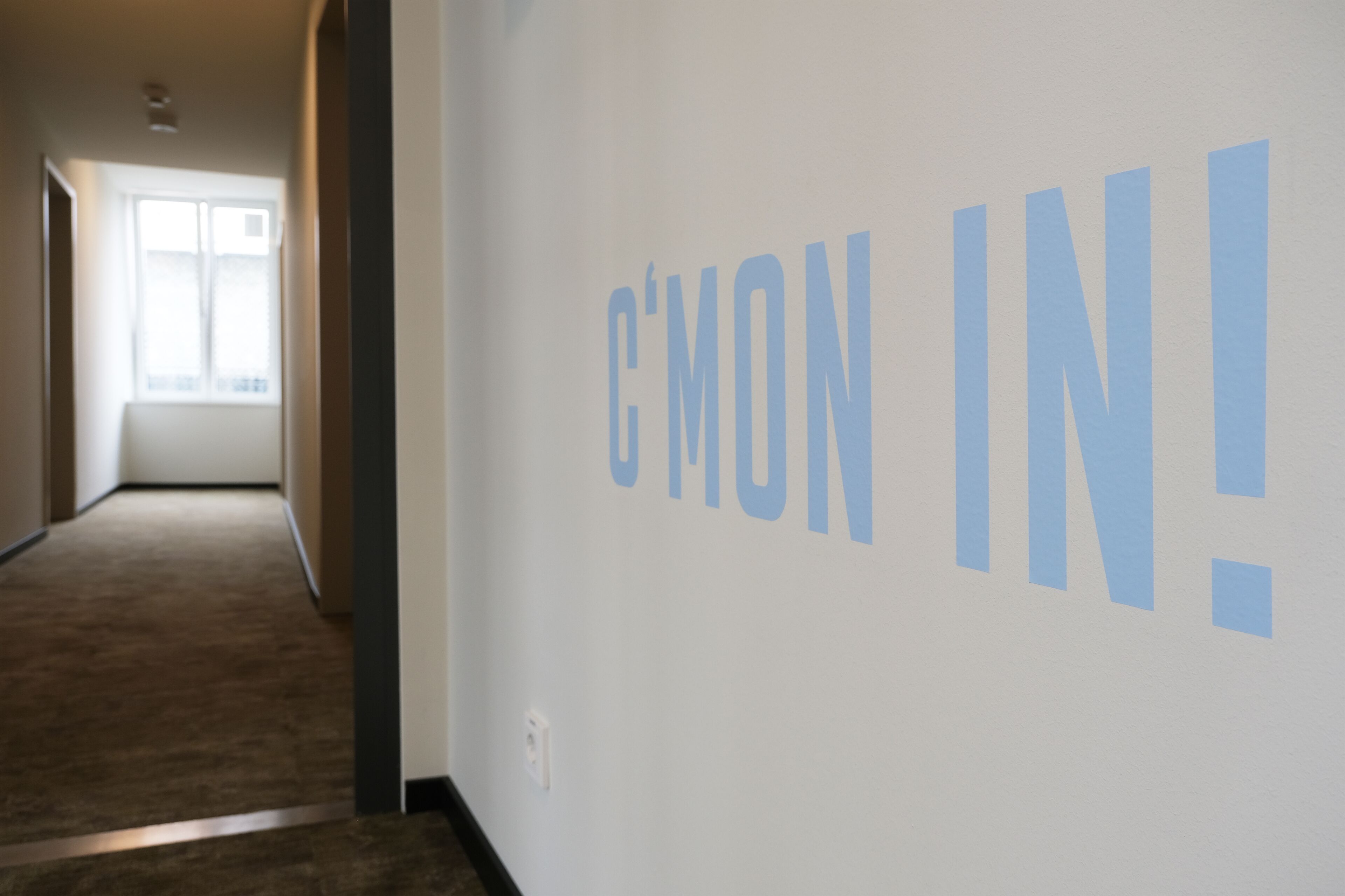 Un pasillo con una pared que muestra el texto 'C'MON IN!' y una ventana al final.