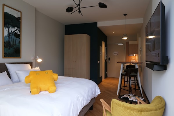 Quarto de hotel com uma cama, um brinquedo de pelúcia amarelo, uma área de refeições e uma televisão fixada na parede.