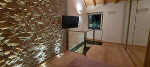 Une pièce avec un mur en pierre, un téléviseur fixé sur un meuble, une fenêtre et un escalier.