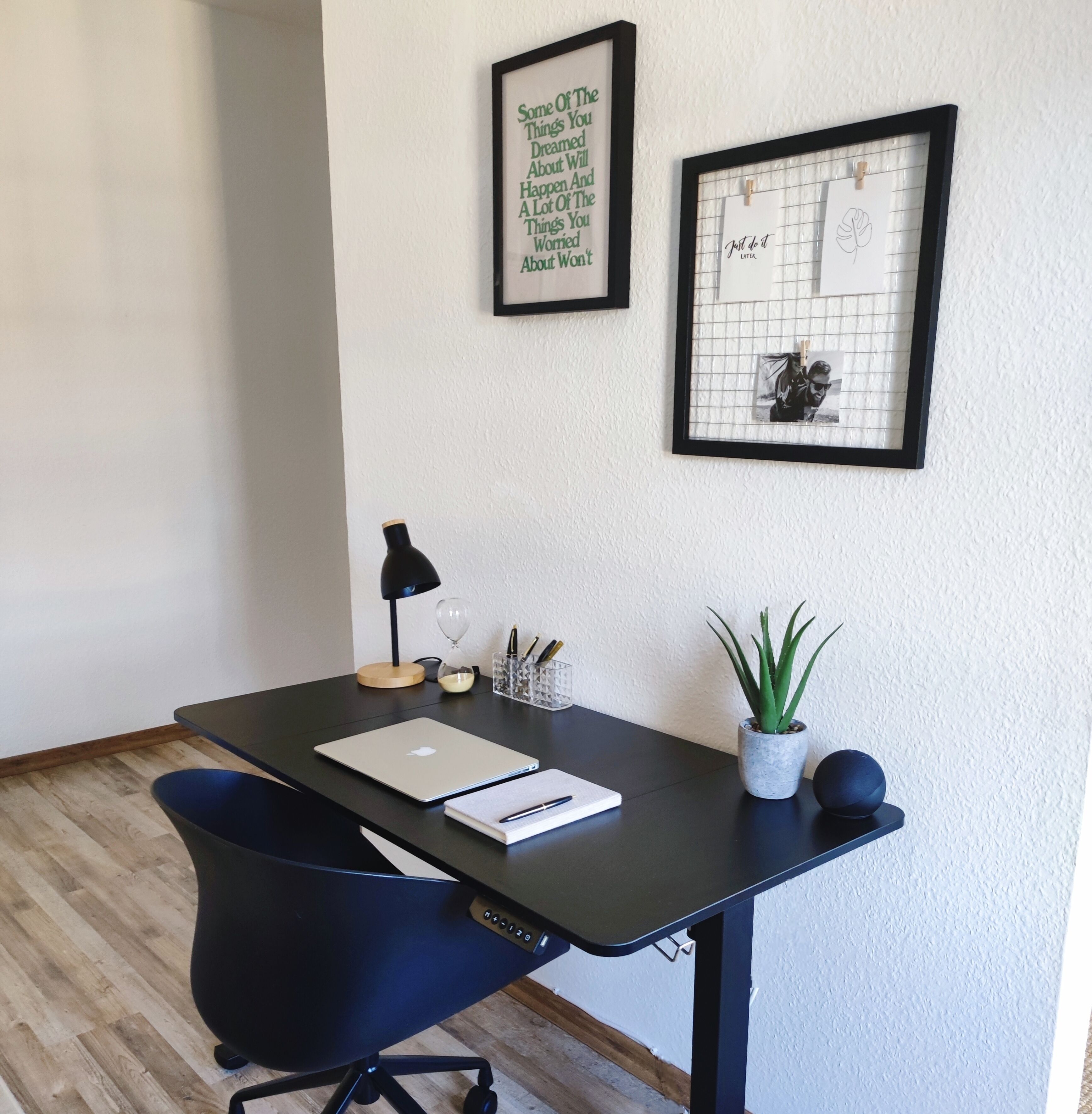 Ein moderner Schreibtisch mit Laptop, Lampe, Topfpflanze und gerahmten Motivationssprüchen an der Wand.