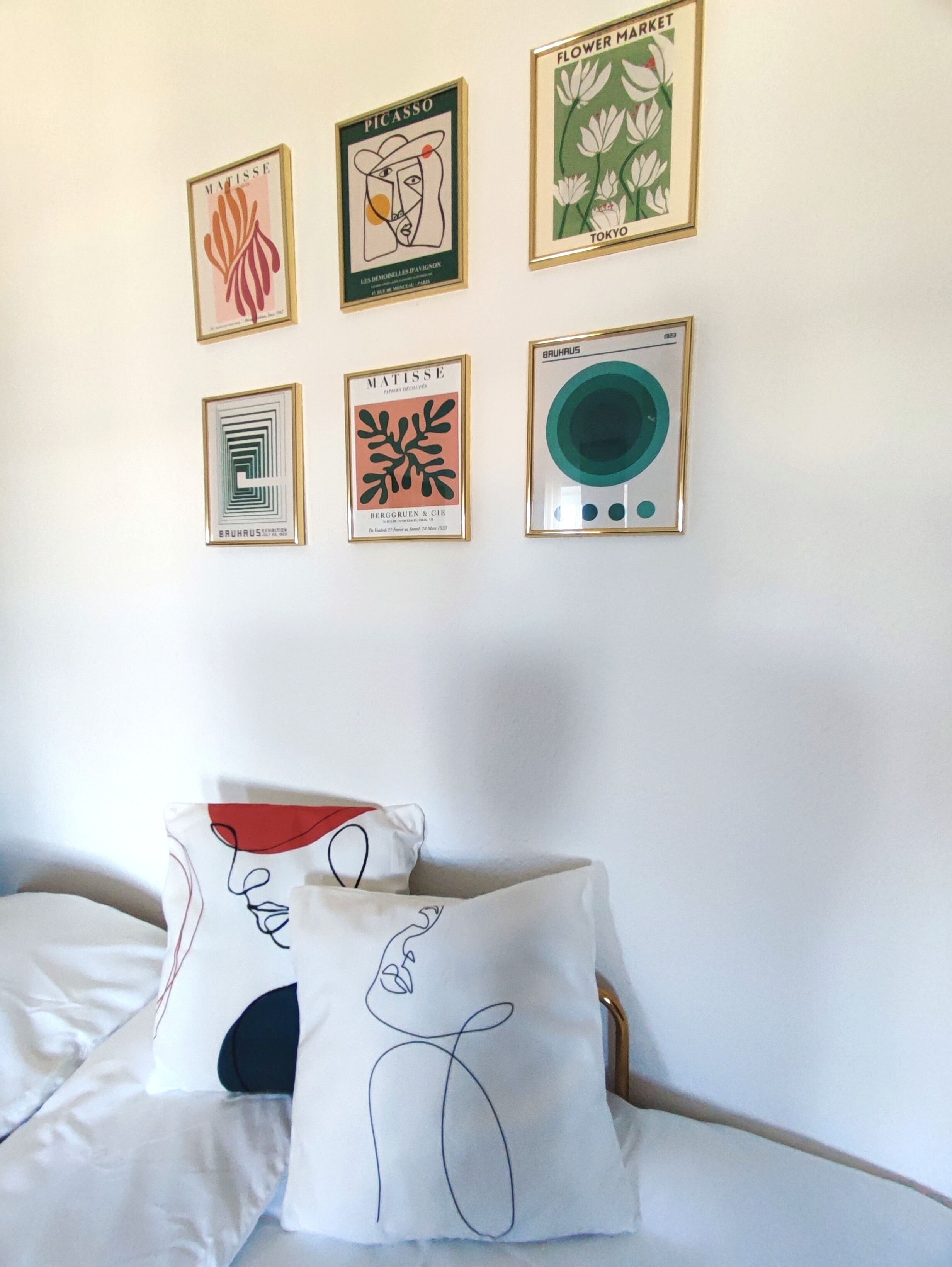A bedroom with a bed, a pillow with abstract art, and four framed artworks on the wall.