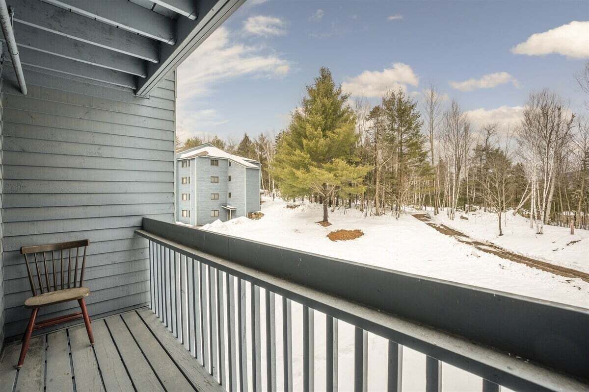 Un balcon avec une chaise en bois, une vue sur un immeuble et un paysage enneigé avec des arbres.