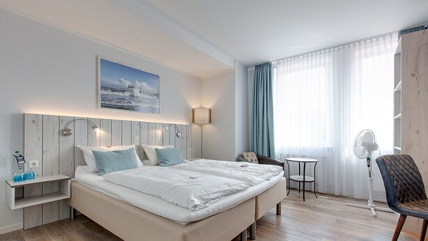 Una habitación de hotel moderna con una cama grande, mesitas de noche, un escritorio, una silla, un ventilador de pie y una ventana con cortinas.