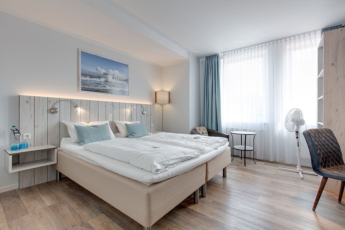 Une chambre d’hôtel moderne avec un grand lit, des tables de chevet, un bureau, une chaise, un ventilateur sur pied et une fenêtre avec des rideaux.