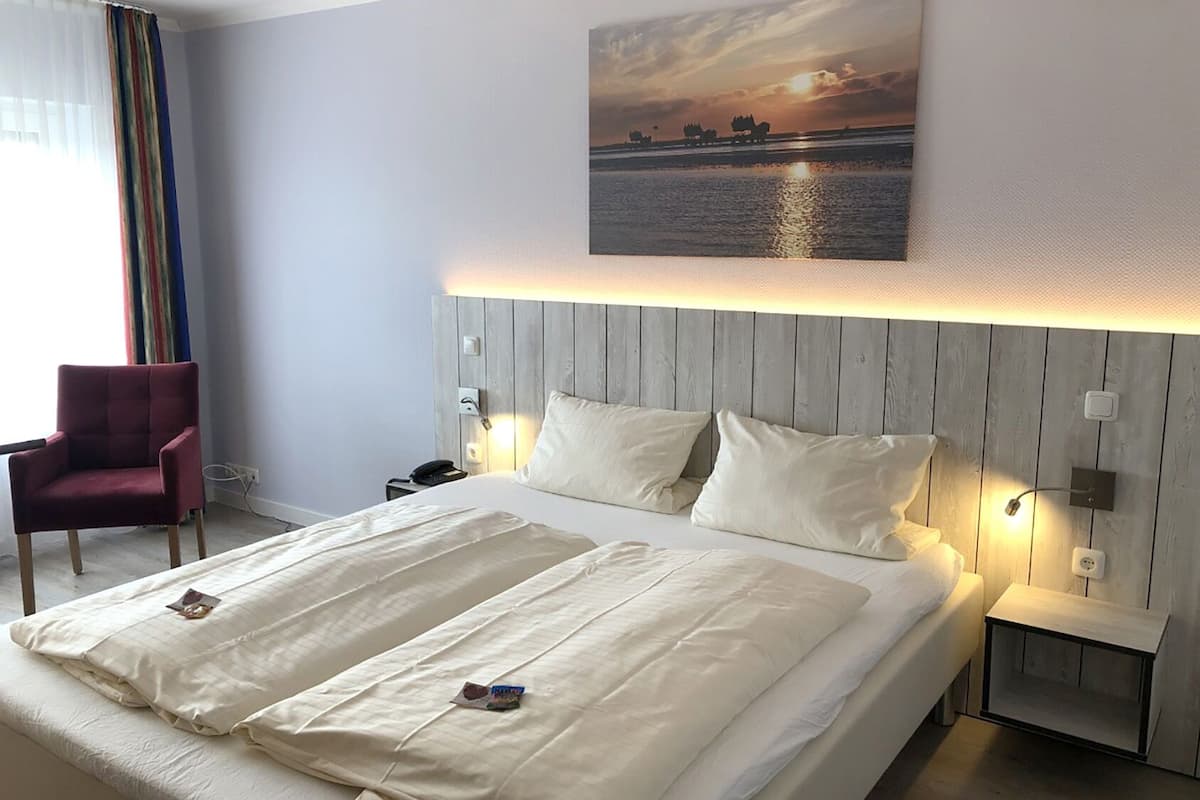Une chambre d’hôtel avec un grand lit, un fauteuil rouge et un tableau représentant un coucher de soleil sur l’eau, accroché au mur.