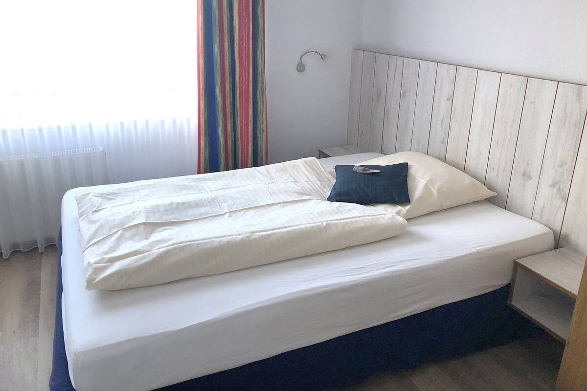 Un lit bien fait, avec du linge de lit blanc et un seul oreiller bleu, une table de chevet en bois et un rideau à rayures, dans une pièce aux murs en bois clair.
