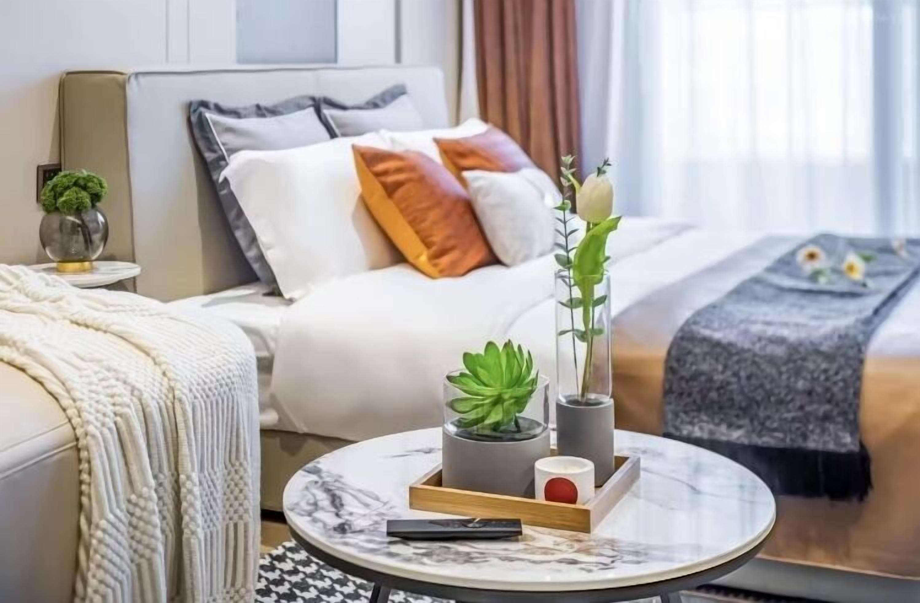 Quarto de hotel moderno com uma cama, uma mesa de centro redonda e uma mesa de cabeceira com uma planta e um vaso.
