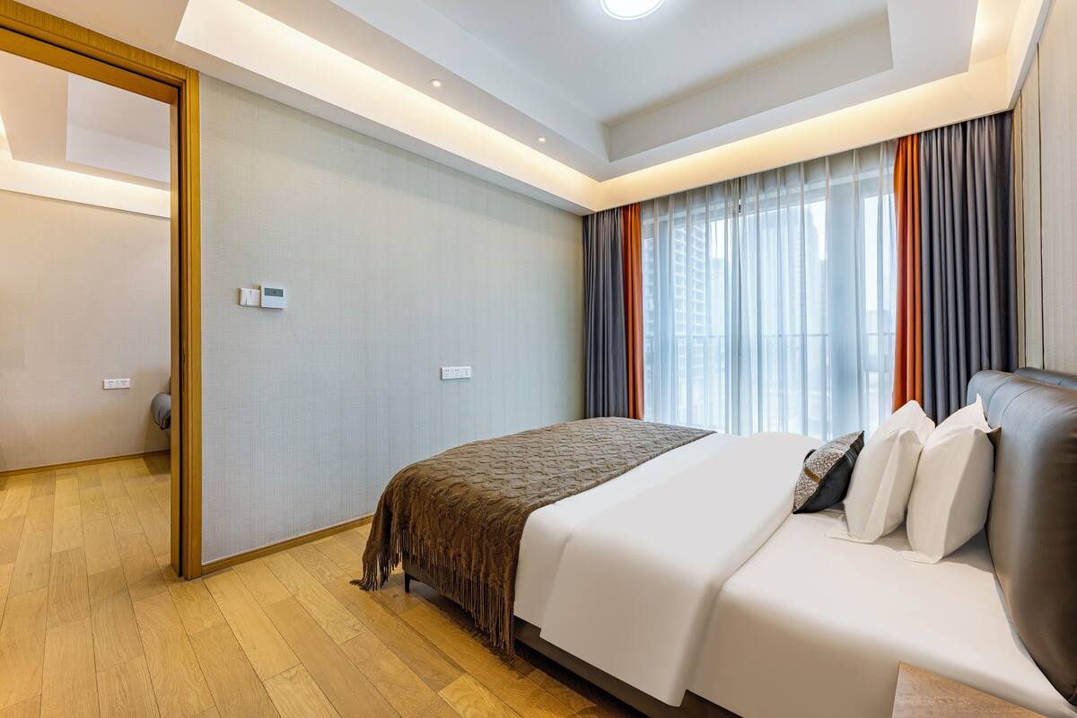A modern hotel room with a large bed, a wooden floor, and a view of the city through large windows.