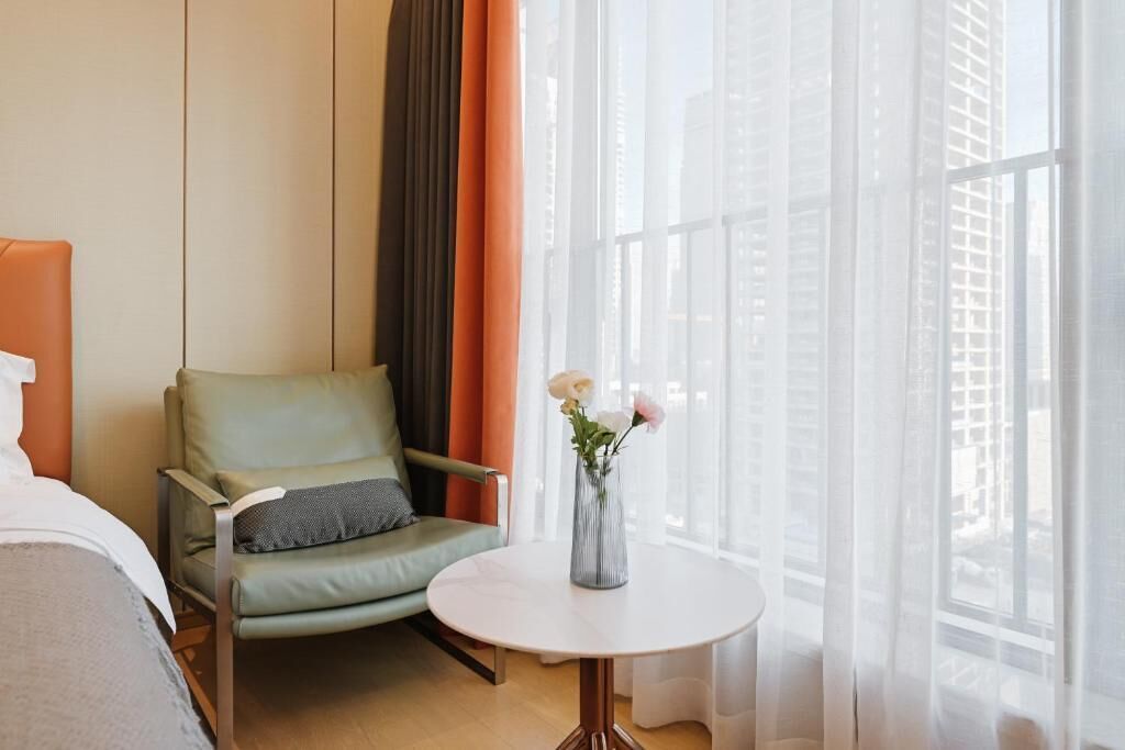 A hotel room with a green armchair, a small round table with a vase of flowers, and a large window with sheer curtains.