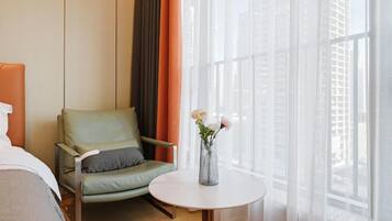 A hotel room with a green armchair, a small round table with a vase of flowers, and a large window with sheer curtains.