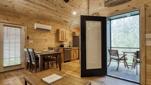 Interior de una cabaña de madera con comedor, cocina y puerta corrediza de vidrio que da a una zona de estar exterior.