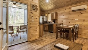 A wooden cabin interior with a dining area, kitchen, and a sliding glass door leading to an outdoor patio.