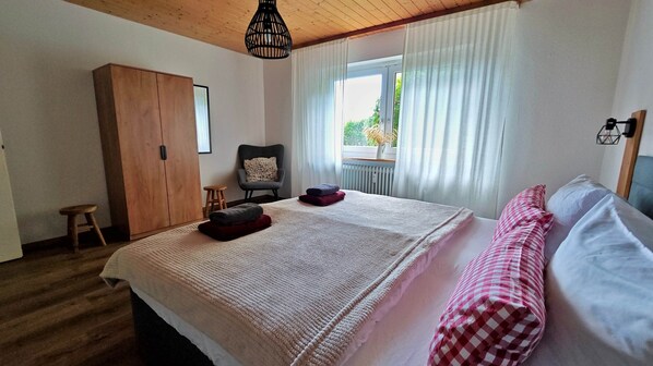Ein Schlafzimmer mit einem großen Bett, einem Holzschrank, einem Stuhl, einem kleinen Hocker und einem Fenster mit Vorhängen.