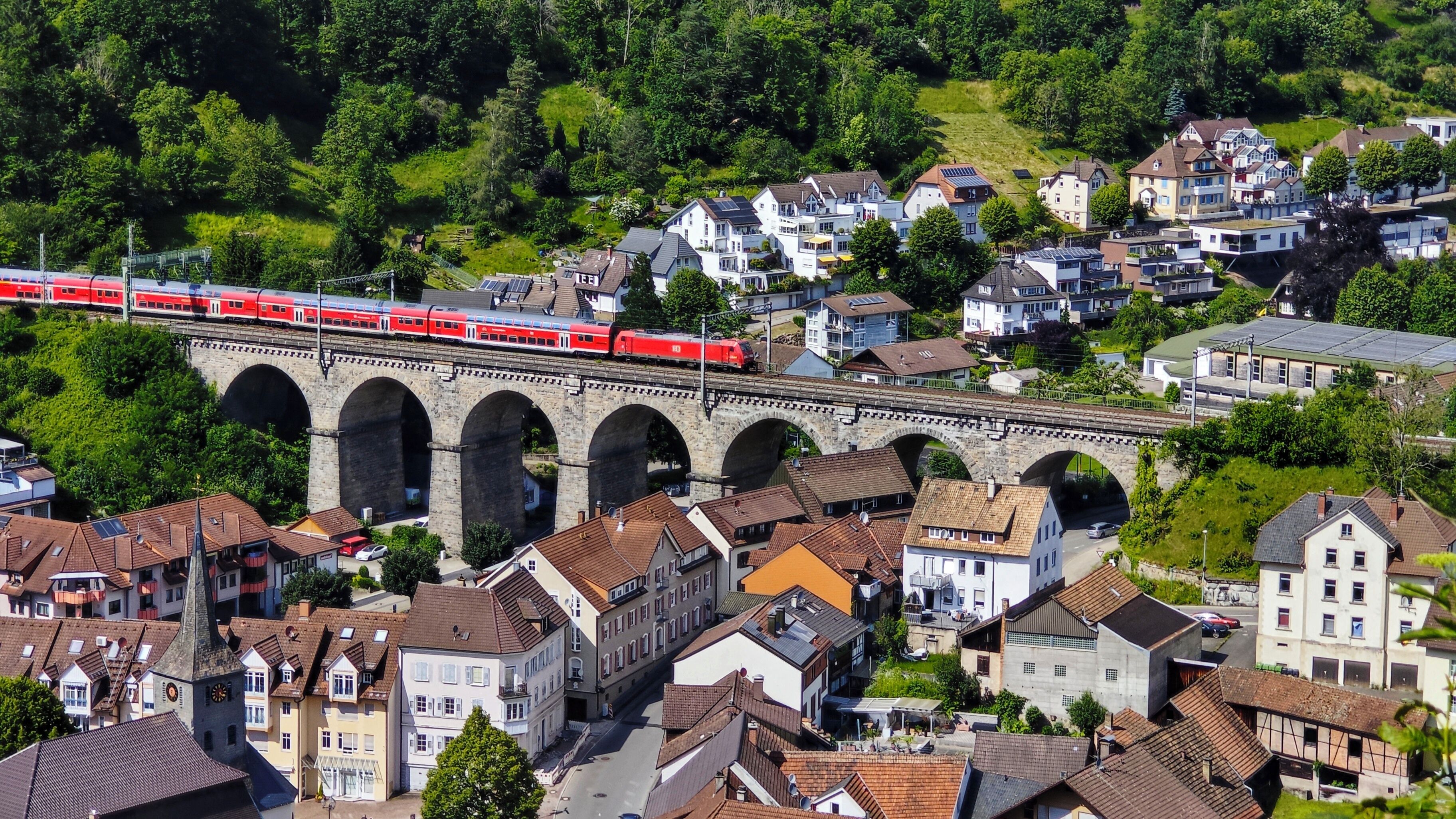Un tren rojo cruzando un puente de piedra sobre una zona residencial con casas y vegetación.
