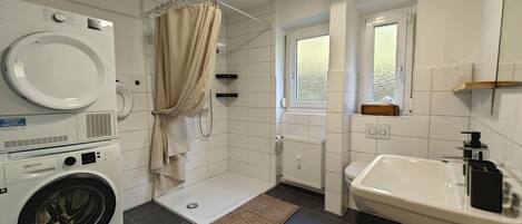 A modern bathroom with a washing machine, a shower, a sink, and a window.