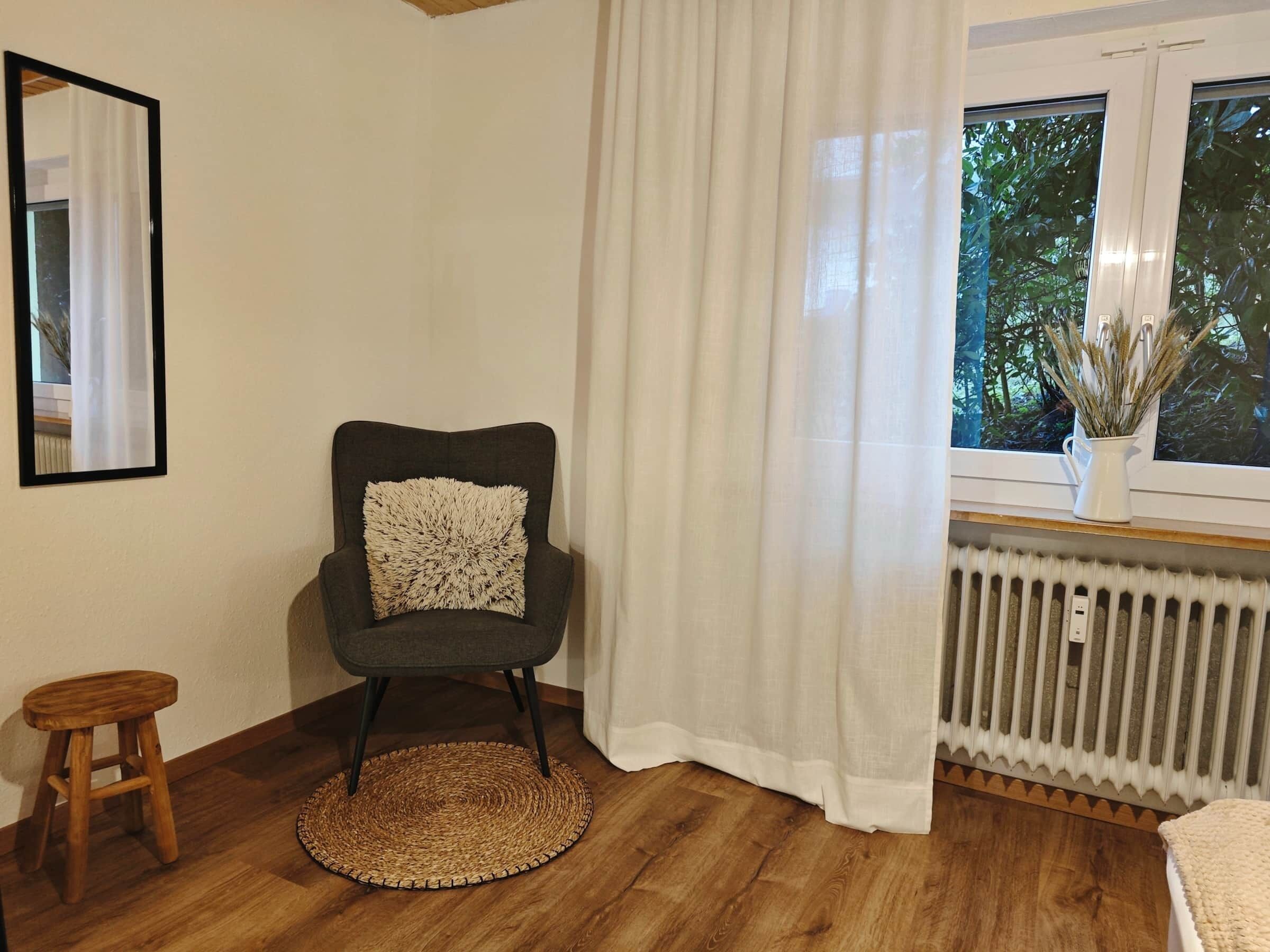 A room with a wooden floor, a dark grey armchair with a textured pillow, a small wooden stool, a round woven rug, a white radiator, and a window with a view of greenery.