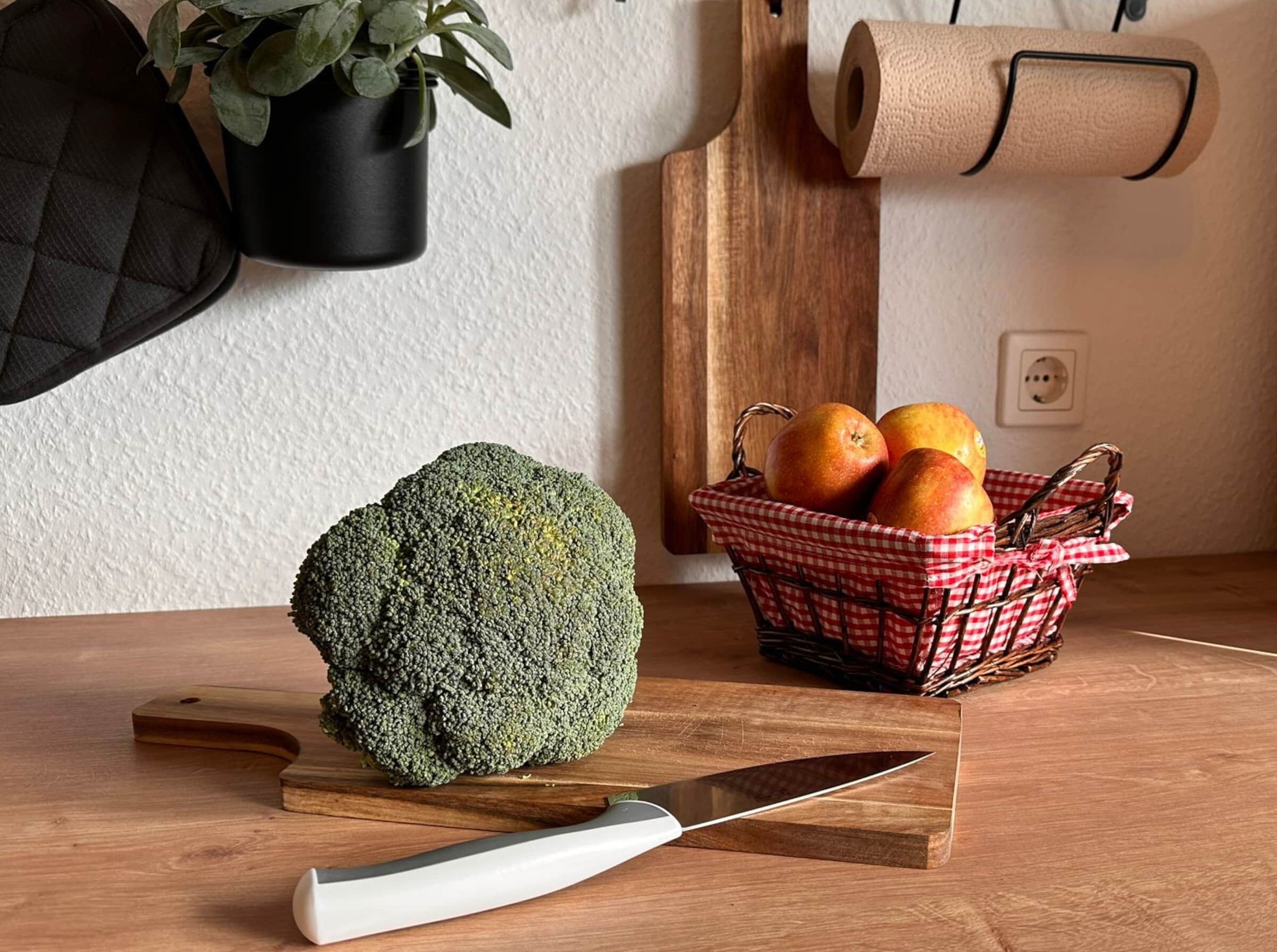 Un taulell de cuina amb un cavolfre gran, un ganivet, una cistella vermella i blanca de cuadros amb pomes i un rotllo de paper de cuina.