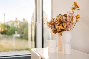 Un florero blanco con flores secas sobre una mesa blanca cerca de una ventana.