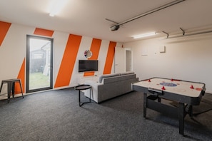 A room with a foosball table, a sofa, a TV, and a window with a view of a house.