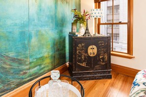 A room with a teal-colored wall, a wooden cabinet with a decorative door, a small table with a crystal decanter, and a window with a view of buildings.