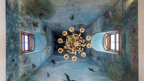 Une pièce dont le plafond est orné d’une fresque représentant des oiseaux et des fleurs, avec deux fenêtres et un lustre.