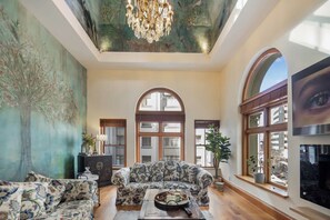 Un salon spacieux avec un grand canapé, une table basse et un lustre. De grandes fenêtres offrent une vue sur l’extérieur.