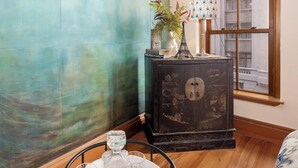 A room with a teal wall, a vintage wooden cabinet with a decorative lamp, and a window with a view of buildings. - The Birds Nest - Central City Loft (Wellington)